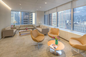 Modern office lounge with large windows showing city skyscrapers. The room has beige armchairs, two sofas, a coffee table with magazines and flowers, and neutral-toned carpet, creating a bright, welcoming atmosphere.