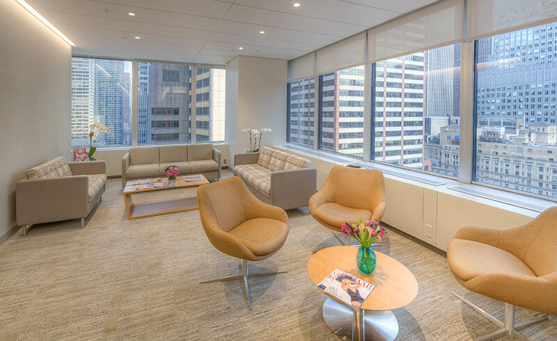 Modern office lounge with large windows showing city skyscrapers. The room has beige armchairs, two sofas, a coffee table with magazines and flowers, and neutral-toned carpet, creating a bright, welcoming atmosphere.