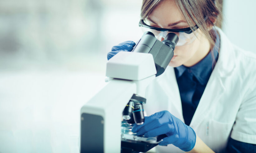 A scientist wearing safety goggles and blue gloves closely examines a sample through a microscope in a laboratory setting.