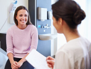 A young woman in a pink sweater smiles while sitting on an exam table, speaking with a healthcare professional holding a clipboard in a medical office.