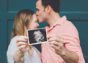 A couple stands close together as the man kisses the woman’s forehead. The woman holds a sonogram photo in front of them, showing an ultrasound image, suggesting they are expecting a baby.