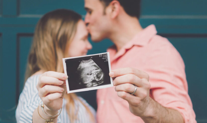 A couple stands close together as the man kisses the woman’s forehead. The woman holds a sonogram photo in front of them, showing an ultrasound image, suggesting they are expecting a baby.