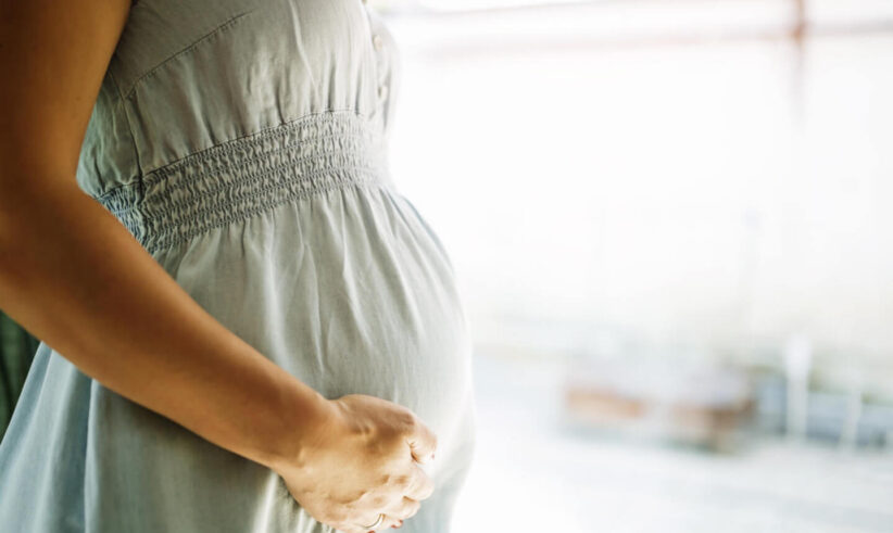 A close-up of a pregnant woman in a light gray dress gently cradling her belly with one hand, standing indoors with soft natural light in the background.