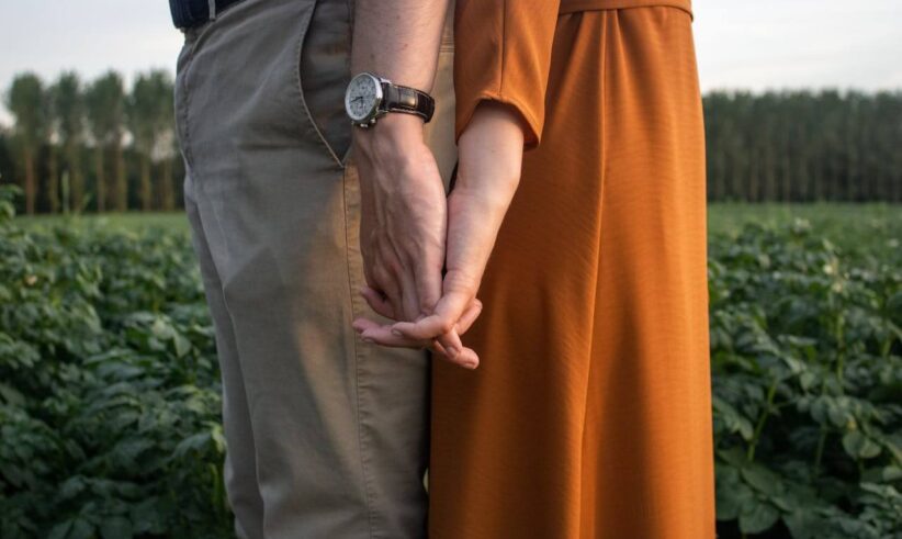 A couple stands close together in a green field, holding hands. One person wears beige pants and a watch, while the other wears an orange dress. Their hands are intertwined, and trees are visible in the background.