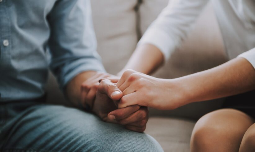 Two people sitting close together on a couch holding hands, one person gently grasping the others hand in a supportive or comforting gesture. Both are wearing casual long-sleeved shirts.
