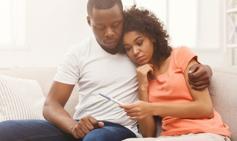 A couple sits closely on a couch, looking intently at a pregnancy test. The man has his arm around the woman, who appears concerned or anxious, resting her head on his shoulder. Both seem deep in thought.