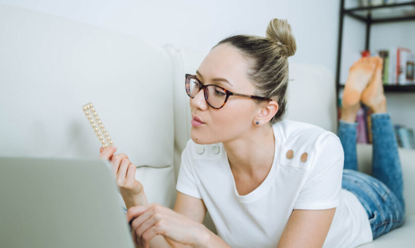 A woman wearing glasses lies on a couch, looking at a pack of pills in one hand while using a laptop with the other hand.
