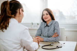 A woman in a light blue shirt sits across from a doctor in a white coat, smiling and talking in a bright office. A stethoscope lies on the desk between them.
