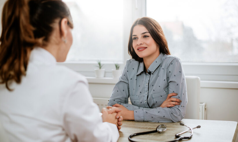 A woman in a light blue shirt sits across from a doctor in a white coat, smiling and talking in a bright office. A stethoscope lies on the desk between them.