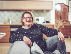 A woman with glasses and shoulder-length hair, wearing a black polka dot top and gray jeans, sits comfortably on a couch in a modern kitchen, smiling at the camera.