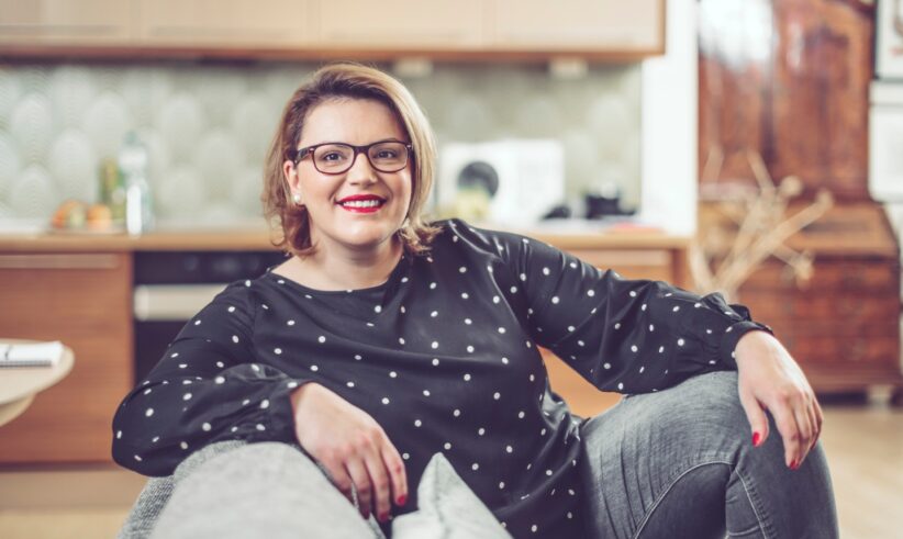 A woman with glasses and shoulder-length hair, wearing a black polka dot top and gray jeans, sits comfortably on a couch in a modern kitchen, smiling at the camera.