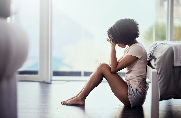 A woman sits on the floor beside a bed, resting her elbows on her knees and her head in her hands, appearing upset or stressed. Sunlight streams through large windows in the background.