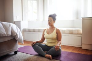 A woman sits cross-legged on a purple yoga mat in a bright room, eyes closed and hands resting on her knees, appearing calm and meditative. Sunlight streams through large windows behind her.