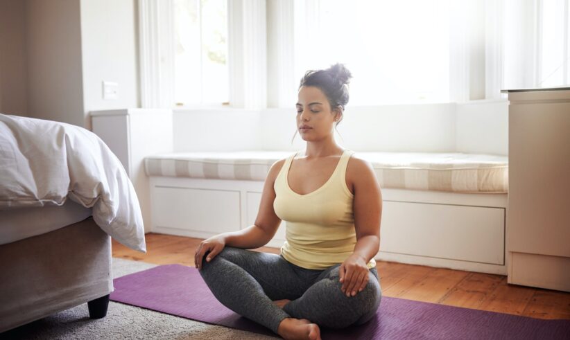 A woman sits cross-legged on a purple yoga mat in a bright room, eyes closed and hands resting on her knees, appearing calm and meditative. Sunlight streams through large windows behind her.