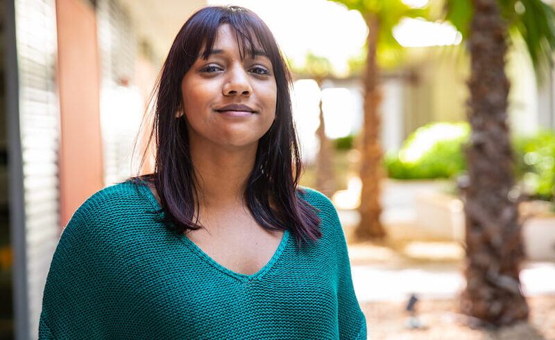 A woman with straight dark hair and bangs, wearing a teal sweater, stands outdoors and smiles softly. The background features palm trees, sunlight, and blurred buildings.
