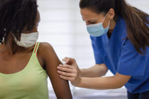 A healthcare worker in blue scrubs and a mask prepares to give a vaccine to a seated woman wearing a mask and a green tank top. The healthcare worker is cleaning the womans upper arm with an alcohol swab.