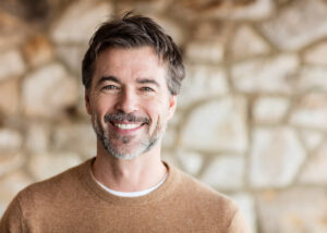 A middle-aged man with short dark hair and a salt-and-pepper beard smiles warmly at the camera. He is wearing a brown sweater and standing in front of a blurred stone wall background.