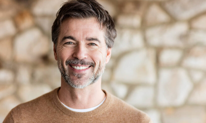 A middle-aged man with short dark hair and a salt-and-pepper beard smiles warmly at the camera. He is wearing a brown sweater and standing in front of a blurred stone wall background.