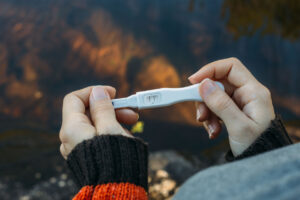A person in a sweater holds a pregnancy test showing a single line, indicating a negative result. The background is blurred with natural outdoor colors.