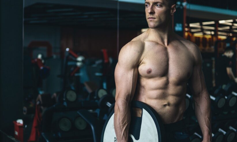 A muscular man stands in a gym holding a large weight plate by his side. He is shirtless, wearing dark workout shorts, and looking to the side. Gym equipment and mirrors are visible in the background.