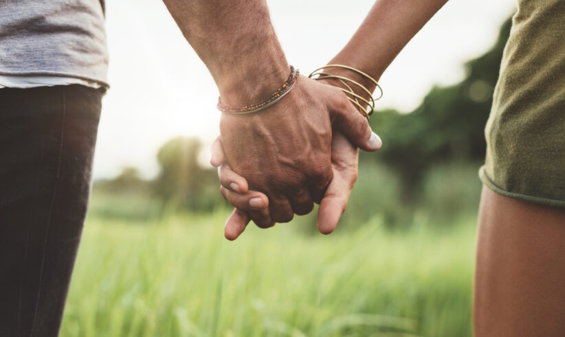 Close-up of two people holding hands outdoors, with one person wearing bracelets. Green grass and trees are visible in the blurred background, and sunlight shines softly between them.