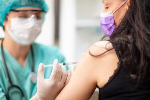 A healthcare worker wearing a mask, face shield, and gloves administers a vaccine injection into the upper arm of a woman who is also wearing a mask.