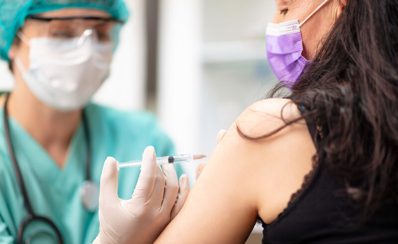 A healthcare worker wearing a mask, face shield, and gloves administers a vaccine injection into the upper arm of a woman who is also wearing a mask.