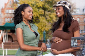 Two women smiling and talking outdoors; one is pregnant and touching her belly, while the other stands nearby with her hand resting on the pregnant womans arm. Trees and buildings are visible in the background.