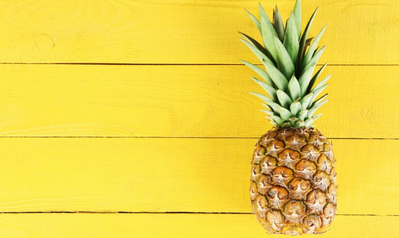 A whole pineapple with green leaves sits on the right side of a bright yellow wooden background.