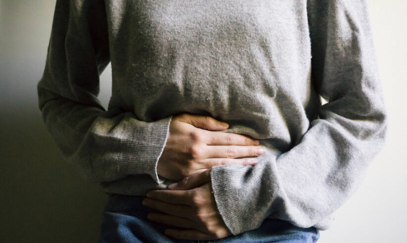 A person in a gray sweater and blue jeans holds their stomach with both hands, suggesting they may have abdominal pain or discomfort.