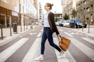 A woman with glasses walks confidently across a city crosswalk, carrying a large brown handbag and wearing a black blazer, white shirt, blue jeans, and white sneakers. Buildings and parked cars line the street.