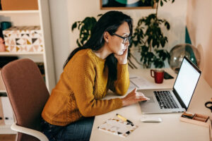 Woman with long dark hair and glasses wearing a mustard sweater sits at a desk, looking intently at a laptop screen. The desk has a red mug, papers, a pen, and a mouse. There are shelves and plants in the background.