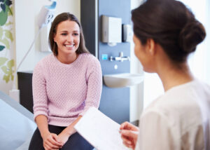 A young woman in a pink sweater smiles while sitting on an exam table, talking to a healthcare professional who is holding a clipboard in a medical office.