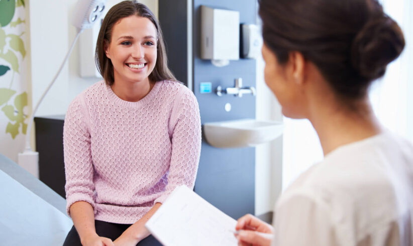A young woman in a pink sweater smiles while sitting on an exam table, talking to a healthcare professional who is holding a clipboard in a medical office.