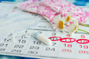 A calendar with several dates circled in red, a white tampon, sanitary pads in pink packaging, and white daisies laid on top, representing menstruation and period tracking.