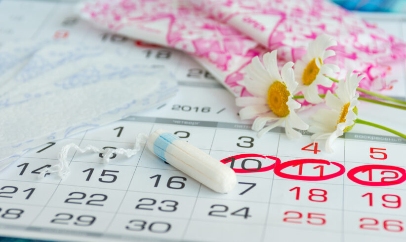 A calendar with several dates circled in red, a white tampon, sanitary pads in pink packaging, and white daisies laid on top, representing menstruation and period tracking.