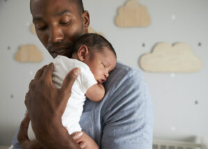 A man gently holds a sleeping newborn baby against his chest in a softly lit nursery with wooden cloud decorations on the wall in the background.