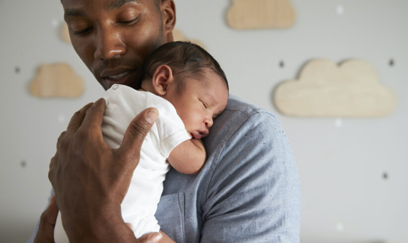 A man gently holds a sleeping newborn baby against his chest in a softly lit nursery with wooden cloud decorations on the wall in the background.