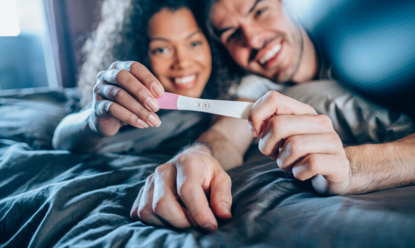 A smiling couple lies on a bed, holding a positive pregnancy test together, expressing excitement and happiness.