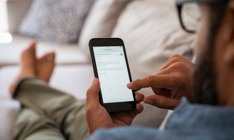 A person reclines on a sofa, using a smartphone with one hand while touching the screen with the other. The phone displays a webpage or app with text and icons. The person wears glasses and casual clothes.