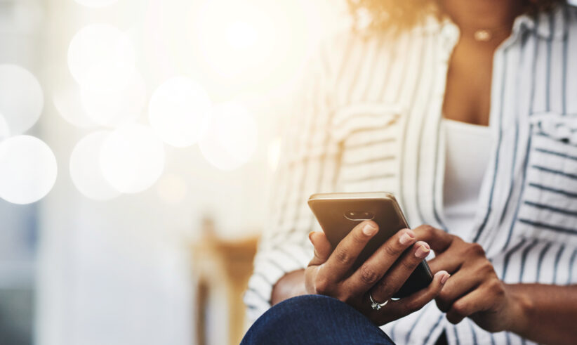 A person wearing a striped shirt sits with crossed legs, holding and using a smartphone. The background is softly blurred with bright, warm lighting and circular bokeh effects.