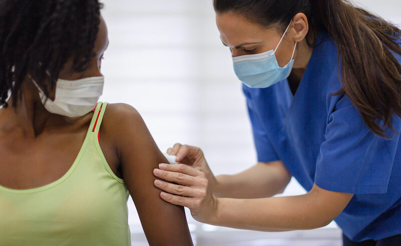 A healthcare worker in blue scrubs and a face mask administers a vaccine to a woman wearing a light green tank top and a mask. The woman looks at her arm as the shot is given.