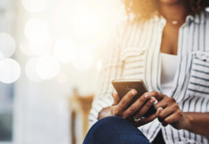 A person wearing a striped shirt sits with legs crossed and uses a smartphone. The background is bright and softly blurred, creating a warm, casual atmosphere.