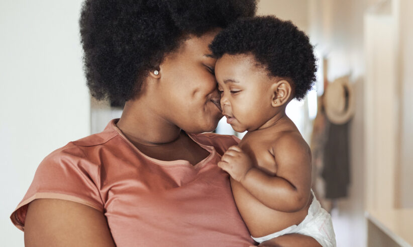 A woman lovingly holds and nuzzles her smiling baby, who is only wearing a diaper. They are indoors, and both share an affectionate moment, their faces close together.