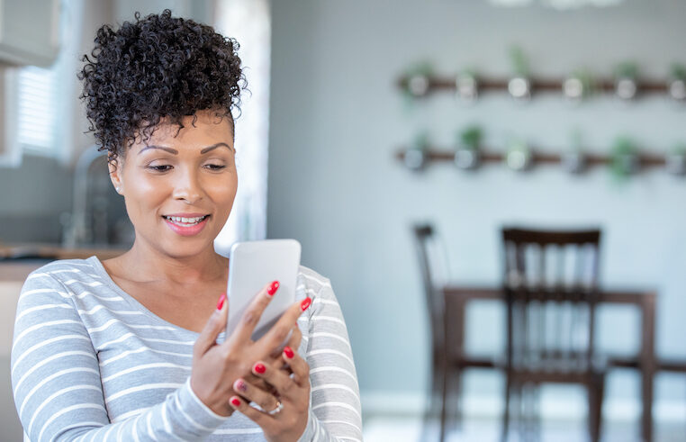 A woman with curly hair and red nails, wearing a gray and white striped shirt, smiles while looking at her smartphone in a bright, modern kitchen with a dining area in the background.