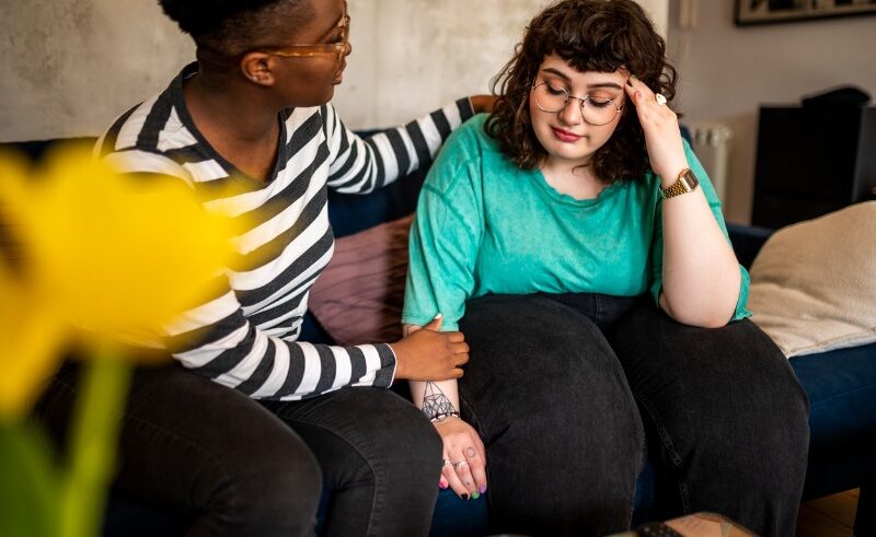Two people sit on a couch; one offers comfort with a hand on the others arm. The person being comforted looks upset, holding their head, while the other shows support. A yellow flower is blurred in the foreground.