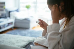 A person in a white bathrobe sits indoors, holding a thermometer and touching their forehead, suggesting they might be checking for a fever. The background shows a living room with soft, natural lighting.