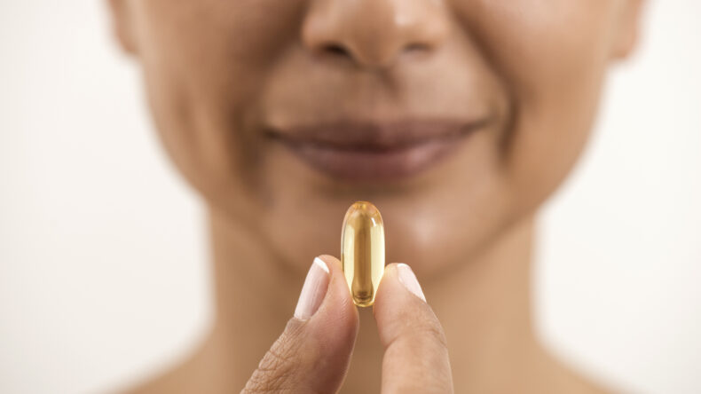 A woman holds a large yellow capsule or soft gel pill between her fingers close to her face, smiling gently with a neutral background.