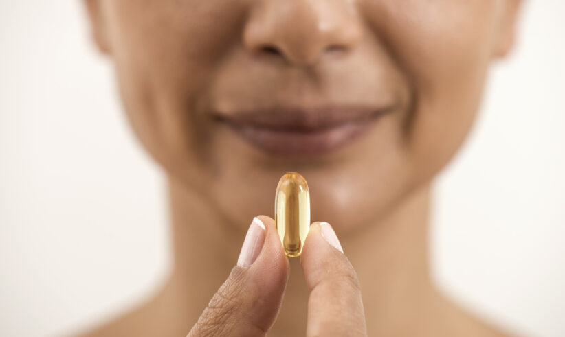 A woman holds a large yellow capsule or soft gel pill between her fingers close to her face, smiling gently with a neutral background.