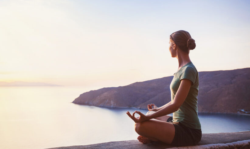 A woman sits cross-legged in a yoga pose on a ledge, facing a calm sea during sunset, with hills in the background and soft sunlight illuminating the scene.